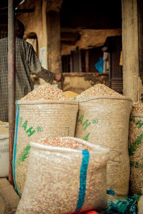 Man holding a smartphone at an open-air grain market surrounded by sacks of grain