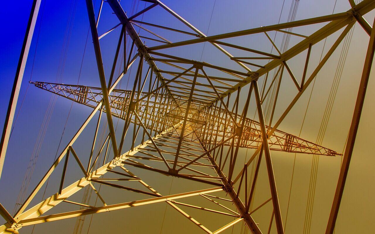 Worker in safety helmet and vest inspecting a steel electricity transmission tower in an open landscape
